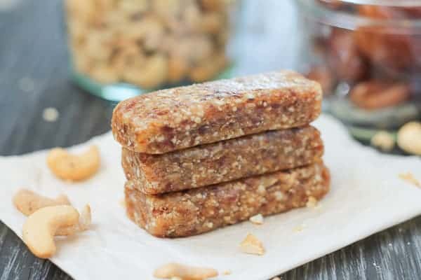 Date bars sitting on a piece of parchment paper with cashews and dates in jars in the background.