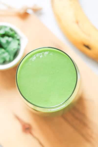 Smoothie in a glass on a light wooden board with a ripe banana and small bowl of greens in the background