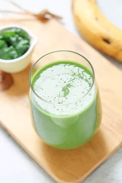 Smoothie in a glass on a light wooden board with a ripe banana and small bowl of greens in the background