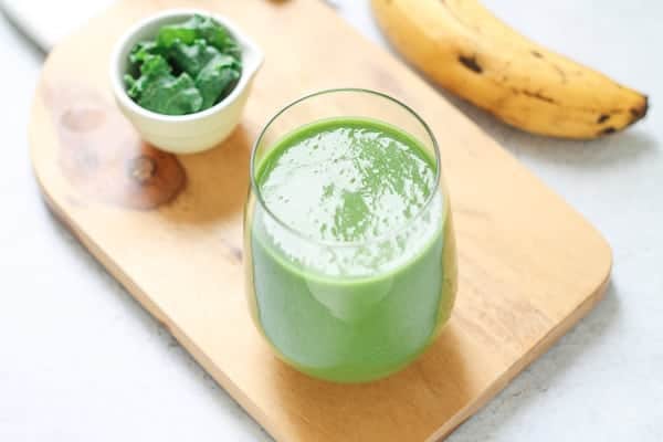 Green smoothie in a glass on a light wooden board with a ripe banana and small bowl of greens in the background