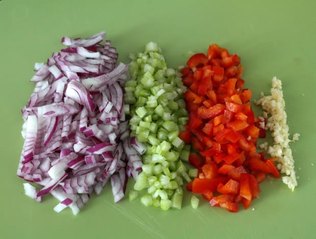 Chopped red onions, celery, bell pepeprs and minced garlic on a green cutting board.