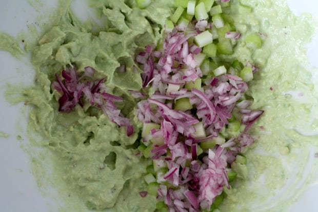 celery and onions being added to a bowl with mashed avocado