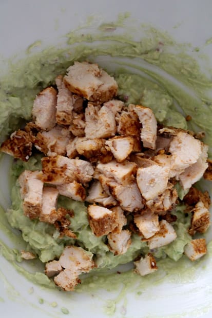 chopped cooked chicken being added to a bowl with mashed avocado