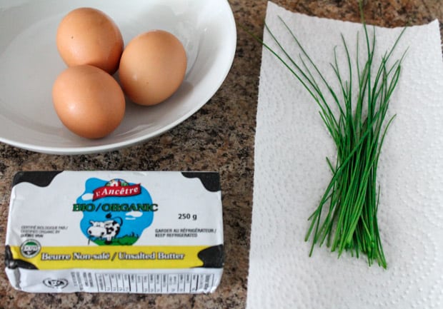 Three eggs in a bowl, fresh chives on a paper towel and a package of butter