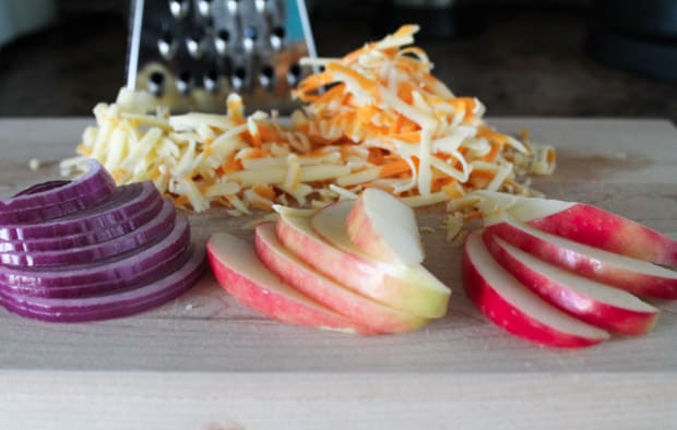 thinly sliced apples and red onions on a cutting board with grated cheese in the background