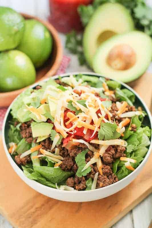 salad topped with spiced beef and shredded cheese with a bowl of fres limes and avocacdo in the background