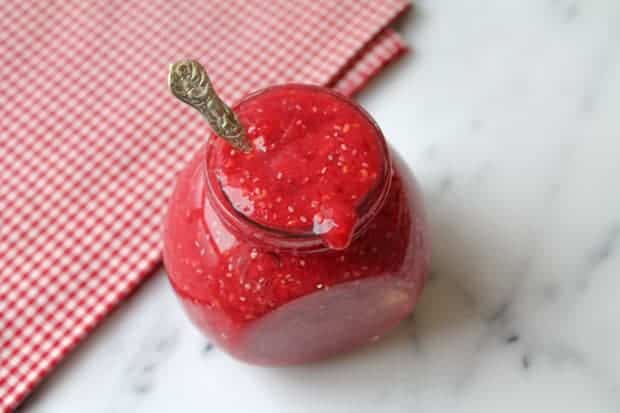 Raspberry chia seed jam in a jar sitting on a a marble surface with a red checkered napkin in the background