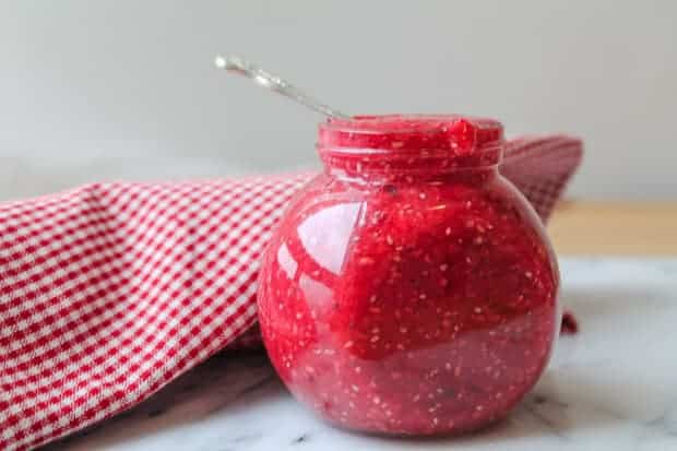 Raspberry chia seed jam in a jar sitting on a a marble surface with a red checkered napkin in the background