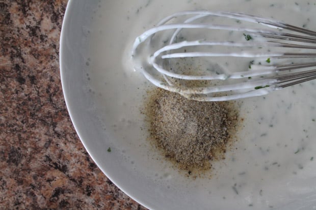 dried seasonings being added to a greek yogurt and milk mixture in a white mixing bowl