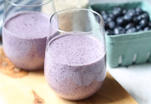 Creamy Blueberry breakfast smoothies in glasses on a wooden board with a jar of oats and basket of blueberries in the background