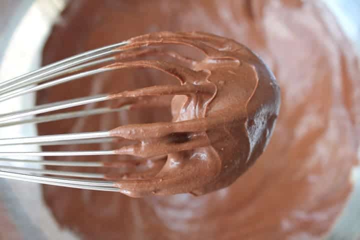 Vegan Chocolate Frosting on a whisk with a bowl full of frosting in the background