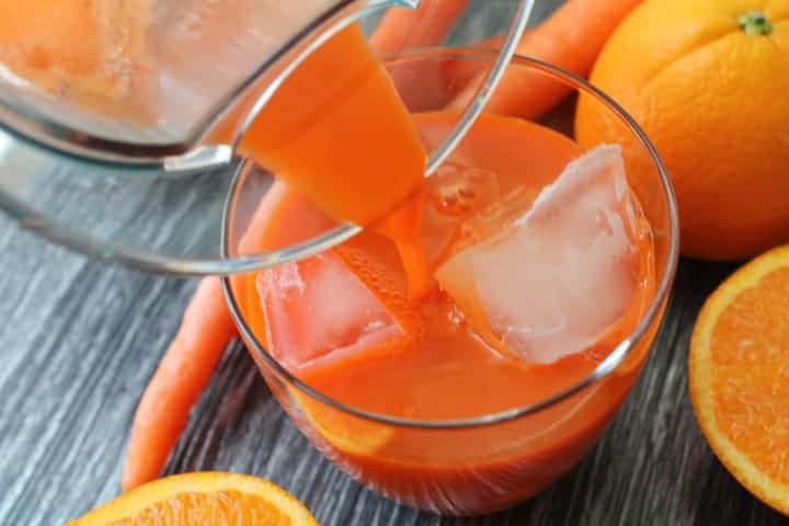 Jugo fresco en un vaso con hielo sobre un fondo de madera rodeado de zanahorias y naranjas en rodajas