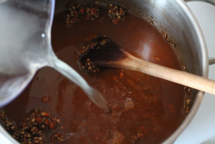 broth being poured into a large soup pot with vegetables and lentils being stirred by wooden spoon.