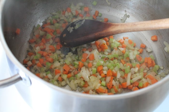 chopped onions, celery and carrots in a large soup pot being stirred by a wooden spoon.