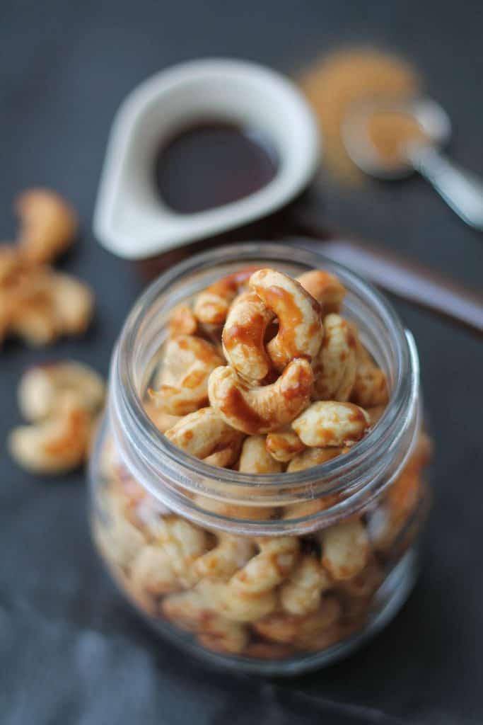 Candied Cashews in a glass jar with a container of maple syrup in the background