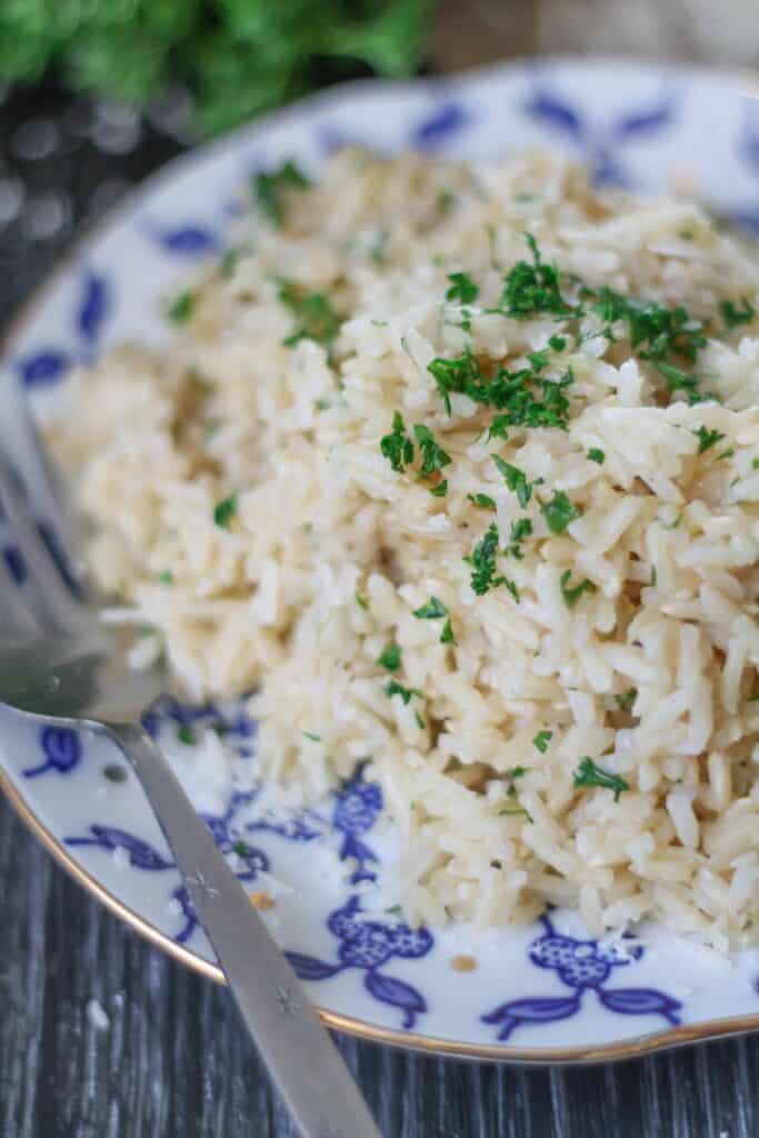 Brown rice sprinkled with parsley on a blue patterned plate with gold trim on a dark wood surface with a bunch of green parsley in the background