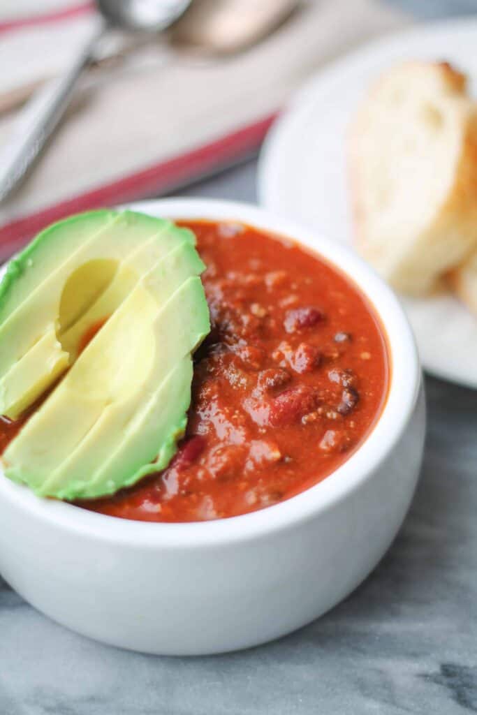 Gluten free Instant Pot Beef Chili topped with fresh avocado in a white bowl on a marble surface with sliced bread in the background