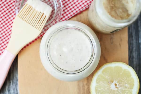 White sauce in a glass jar on a wooden cutting board with a basting brush, jar of horseradish and a cut lemon in the background