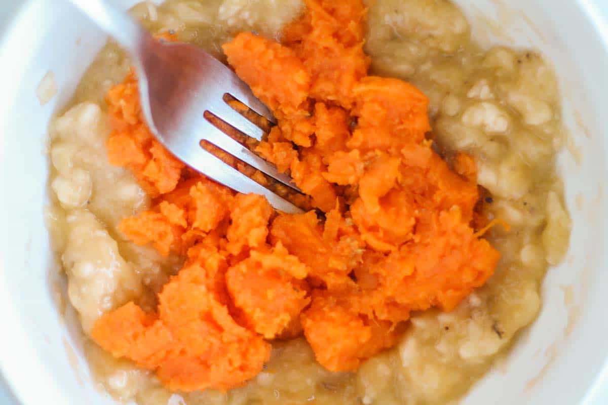 a bowl with cooked sweet potatoes being mashed with bananas by a fork