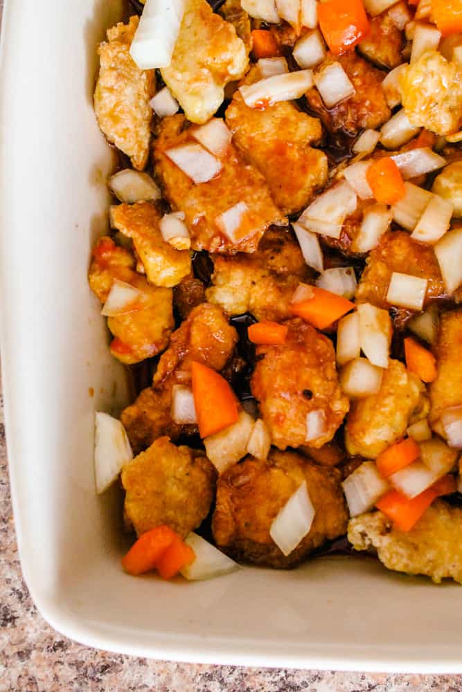 chicken pieces with breading sprinkled with chopped onions and carrots in a casserole dish.