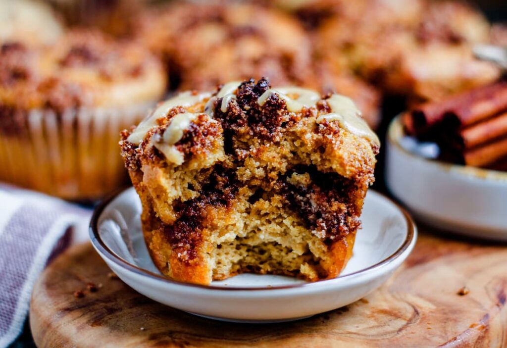 A close-up of a cinnamon muffin with a bite taken out of it, revealing moist, crumbly texture and swirls of cinnamon. It is placed on a small plate, with more muffins and some cinnamon sticks visible in the background.