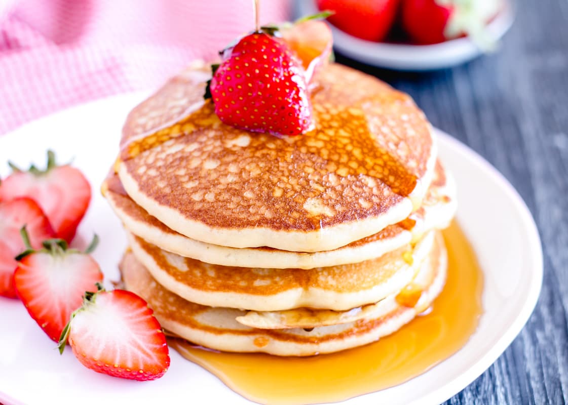 A plate of gluten free pancakes drizzled with maple syrup and garnished with strawberries.
