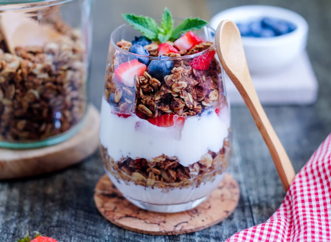 A parfait glass of gluten free granola, fresh fruit, and yogurt with a spoon next to it.