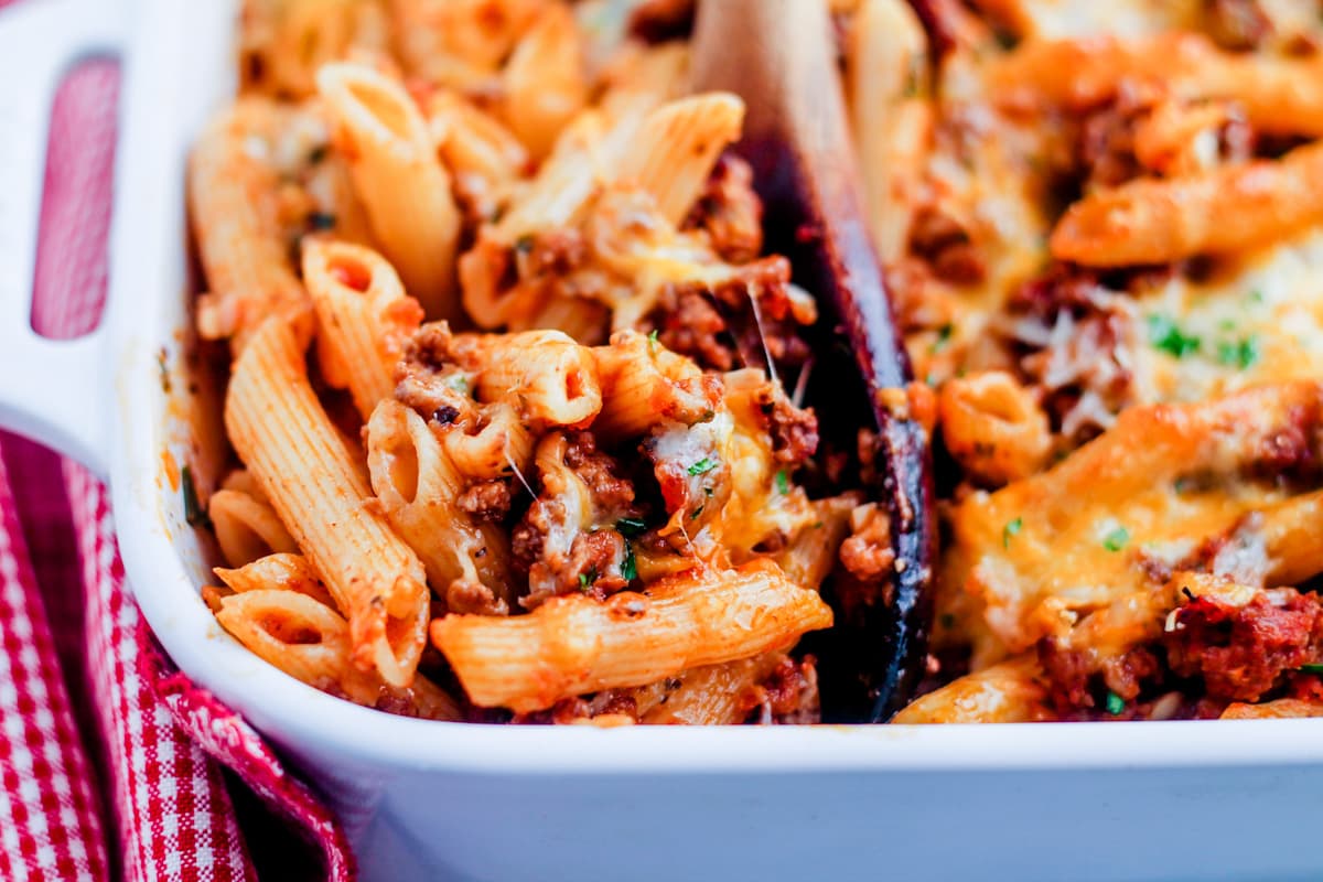 A wooden spoon scooping penne pasta bake from a casserole dish.