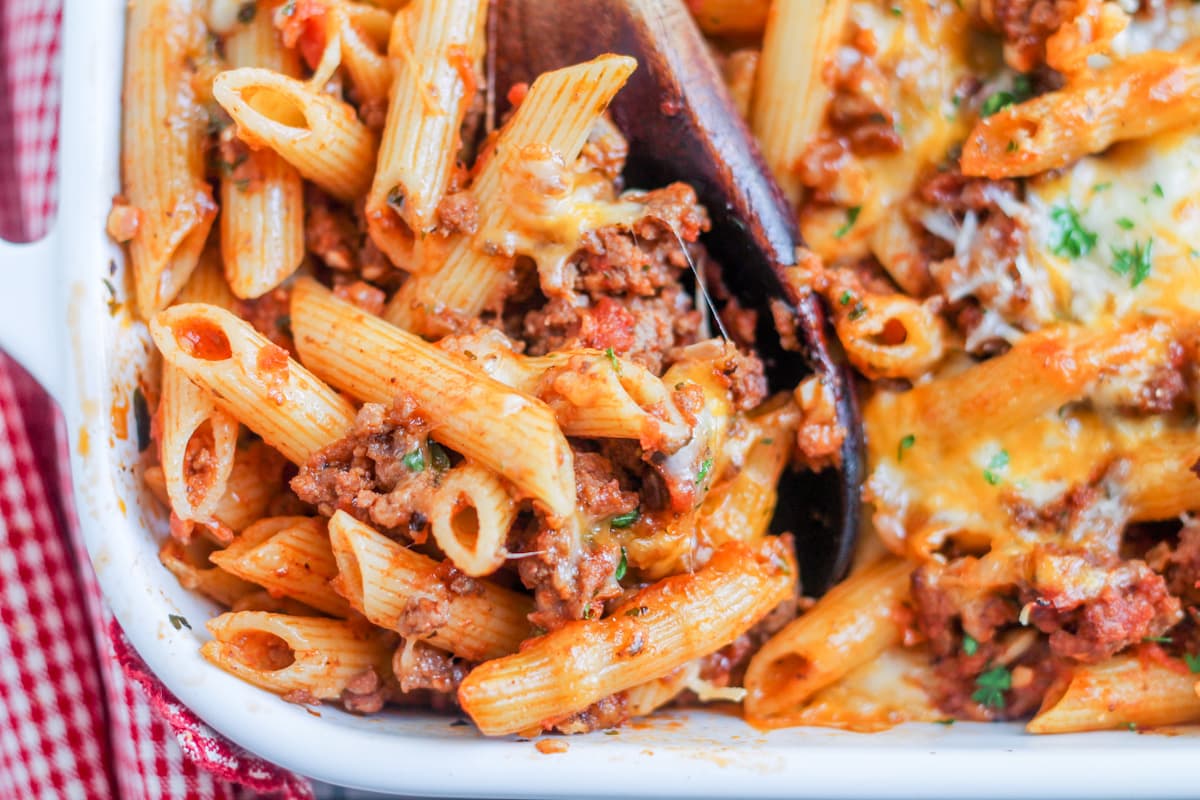 A wooden spoon scooping penne pasta bake from a casserole dish.