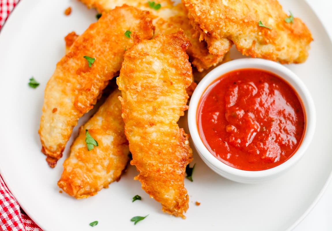 A plate of baked parmesan chicken tenders with a dish of dipping sauce.