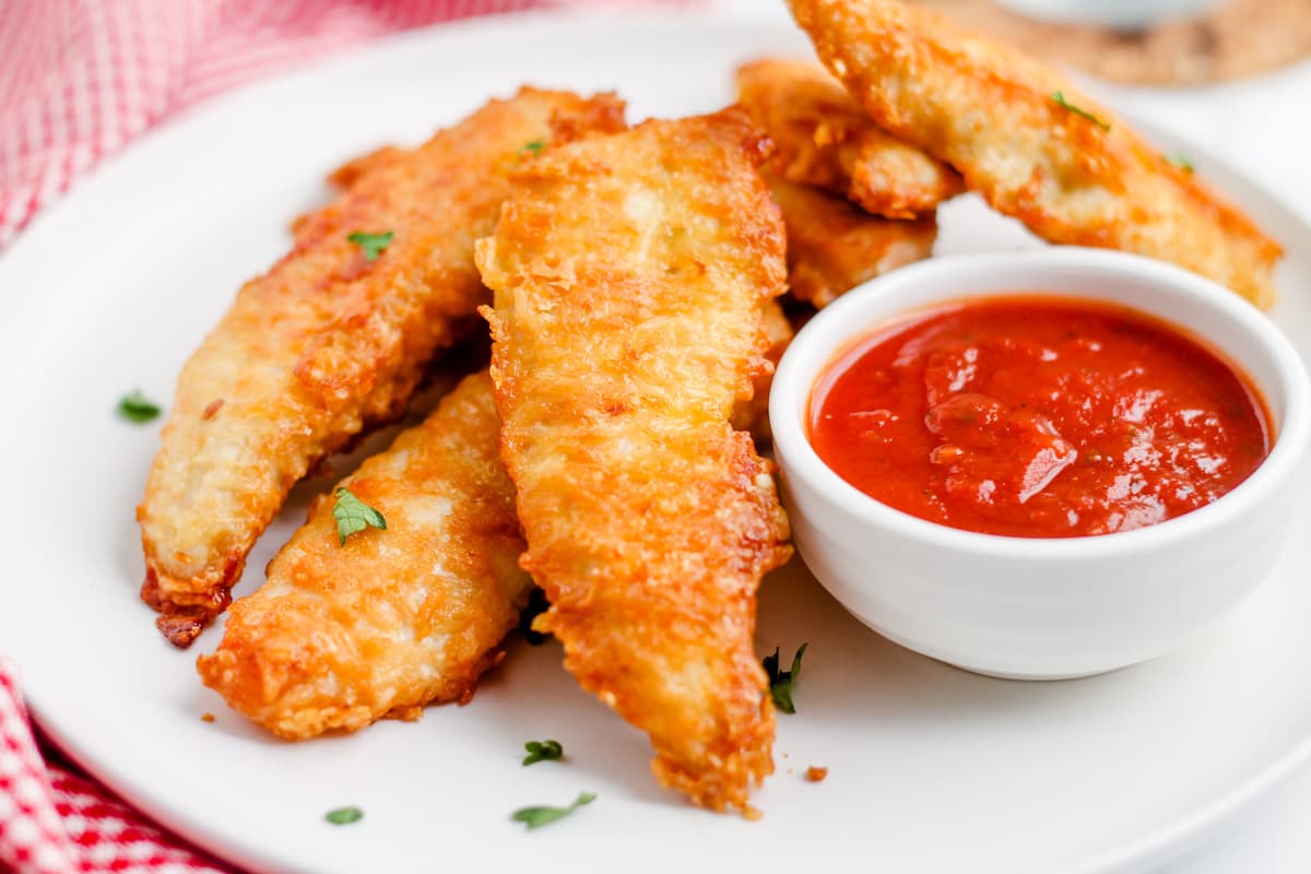 A plate of baked parmesan chicken tenders with a dish of dipping sauce.