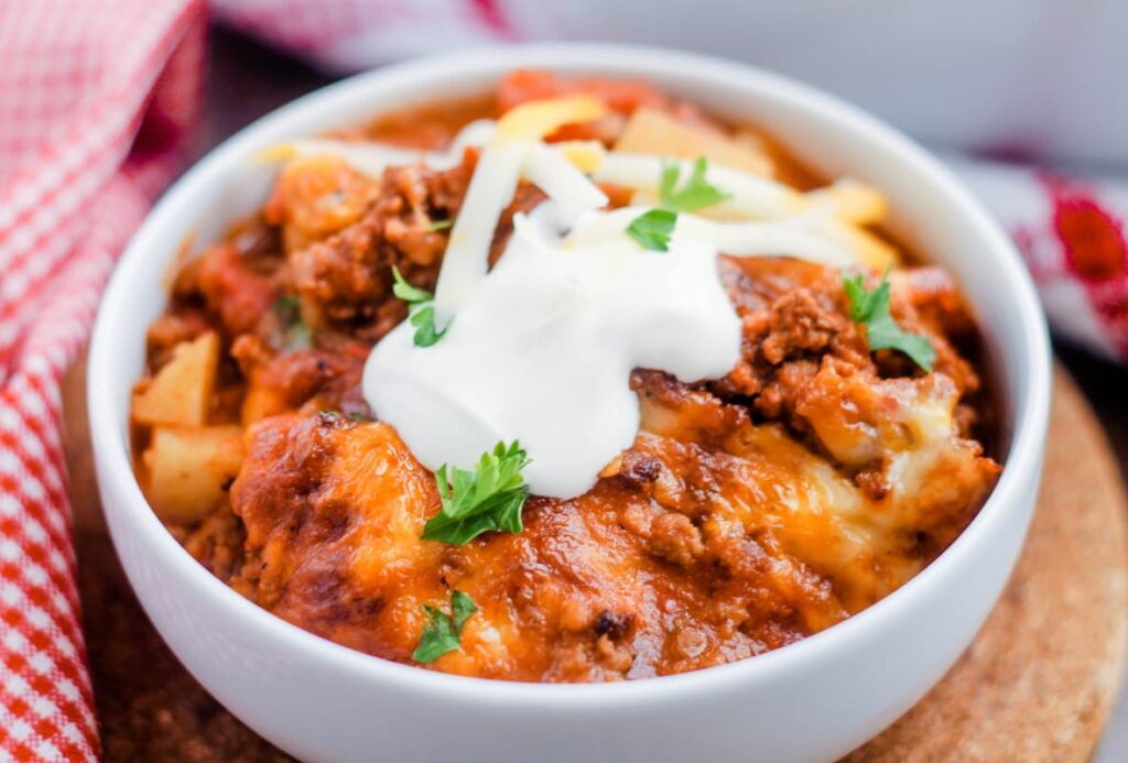 A white bowl filled with cheesy chili, topped with a dollop of sour cream and garnished with fresh parsley. The dish includes chunks of vegetables and ground meat, with a checkered red and white cloth partially visible in the background.