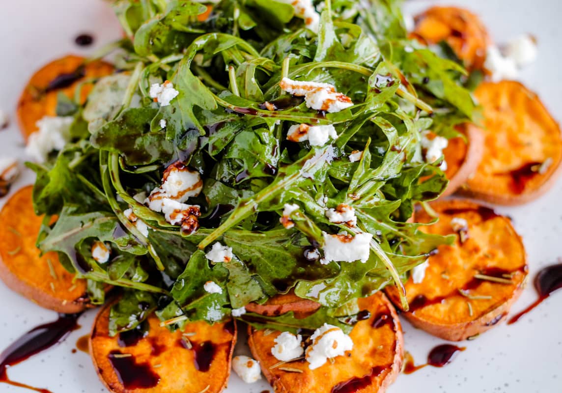 A plate of arugula salad topped with crumbled goat cheese served on top of roasted sweet potatoes and drizzled with balsamic glaze