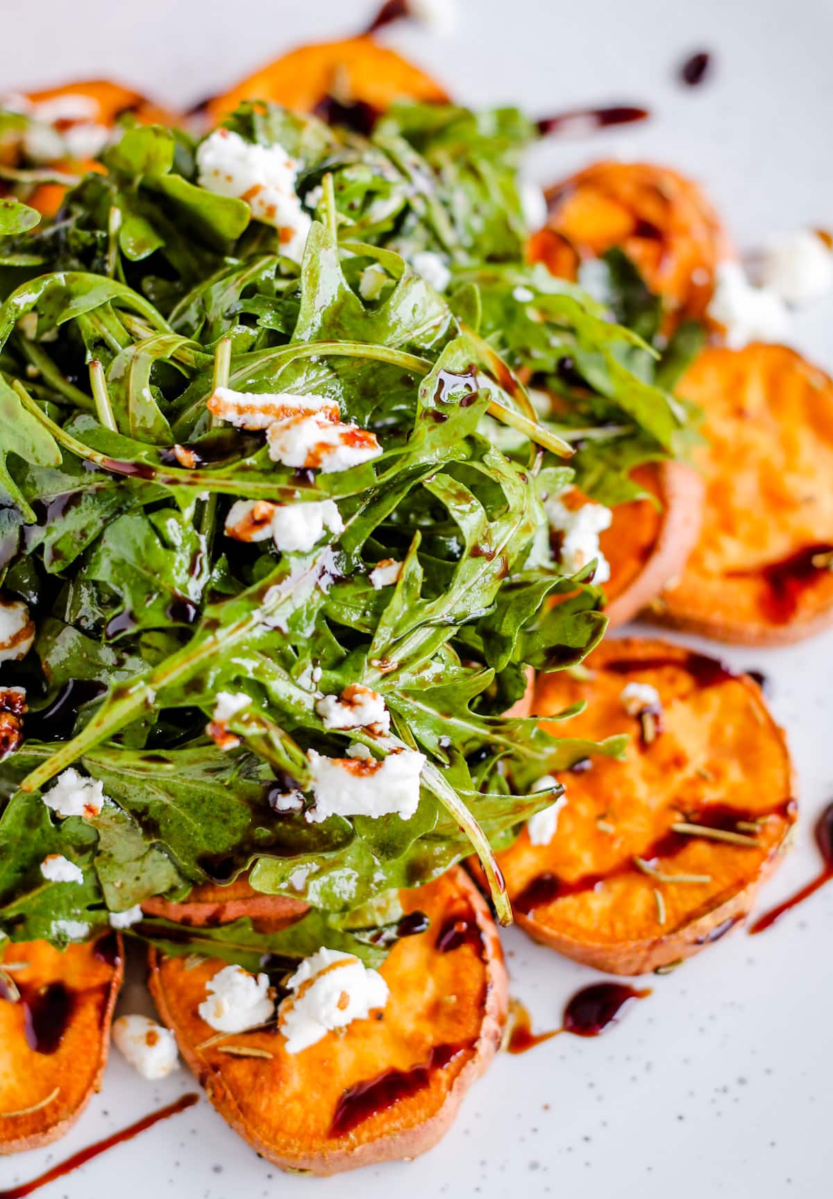A plate with arugula salad topped with crumbled goat cheese served on top of roasted sweet potatoes and drizzled with balsamic glaze