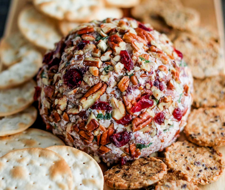 A cranberry pecan cheese ball with crackers on a wooden board.
