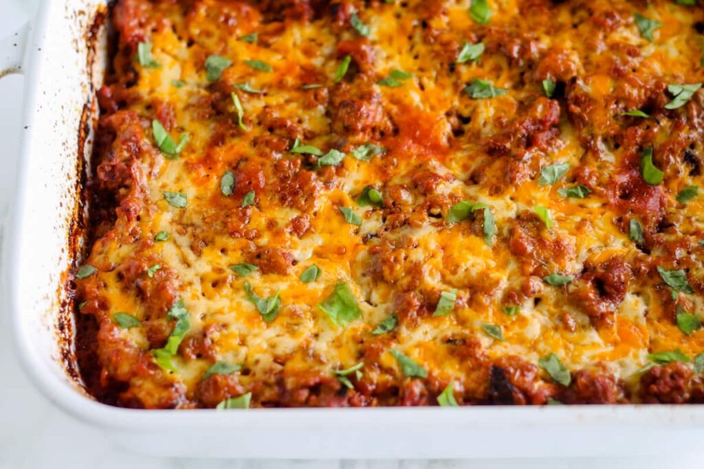 A close-up of a casserole dish filled with baked lasagna. The top layer is golden brown with melted cheese and visible meat sauce. Fresh green herbs are sprinkled on top, adding a pop of color to the dish.