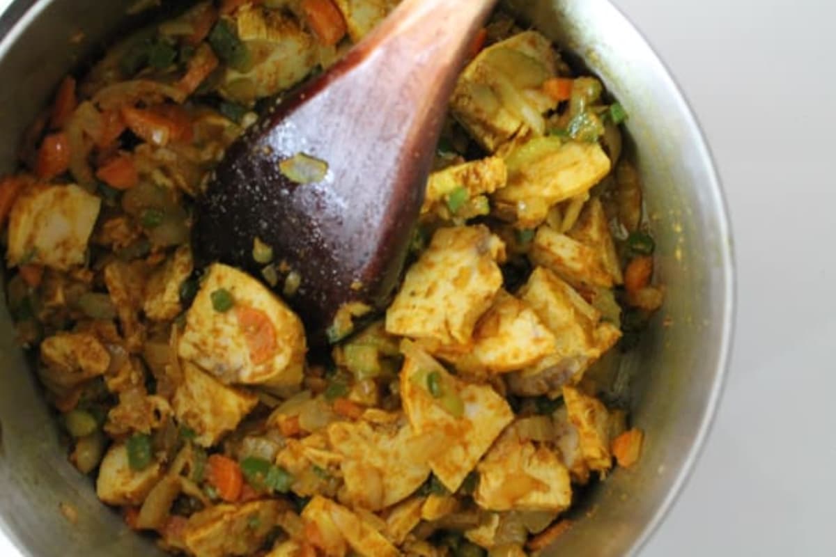 Cooked chicken being stirred into a saucepan with cooked veggies.