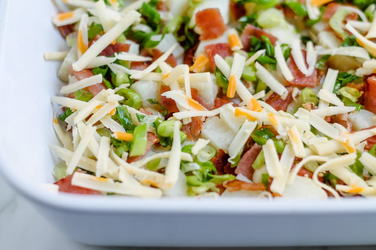 Green onions and cheese sprinkled on potatoes and bacon in a baking dish.